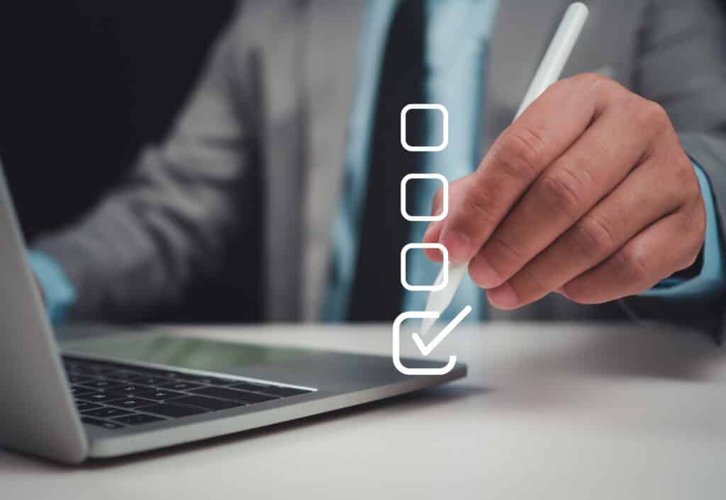 Main cochant une case sur une liste numerique a l'ecran avec un stylet, pres d'un ordinateur portable. Completion de tache en ligne.