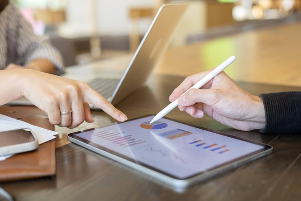 Deux personnes collaborent sur une tablette montrant des graphiques et un diagramme, l'une pointant, l'autre utilisant un stylet. Analyse de données.