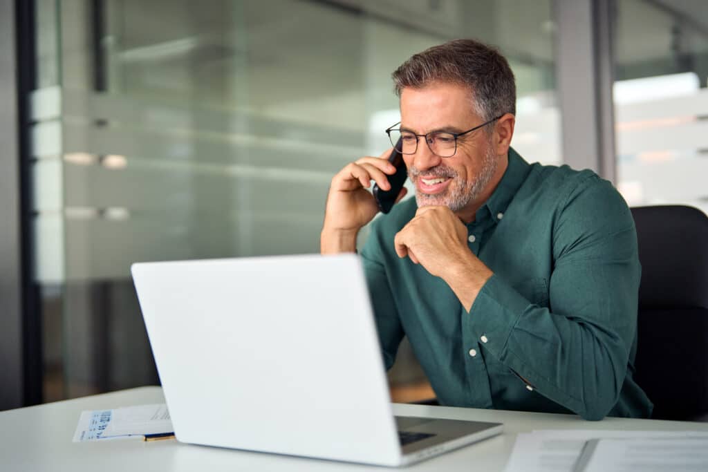 Homme souriant en chemise verte, parlant au téléphone portable, travaillant sur un ordinateur portable au bureau. Heureux et concentré.