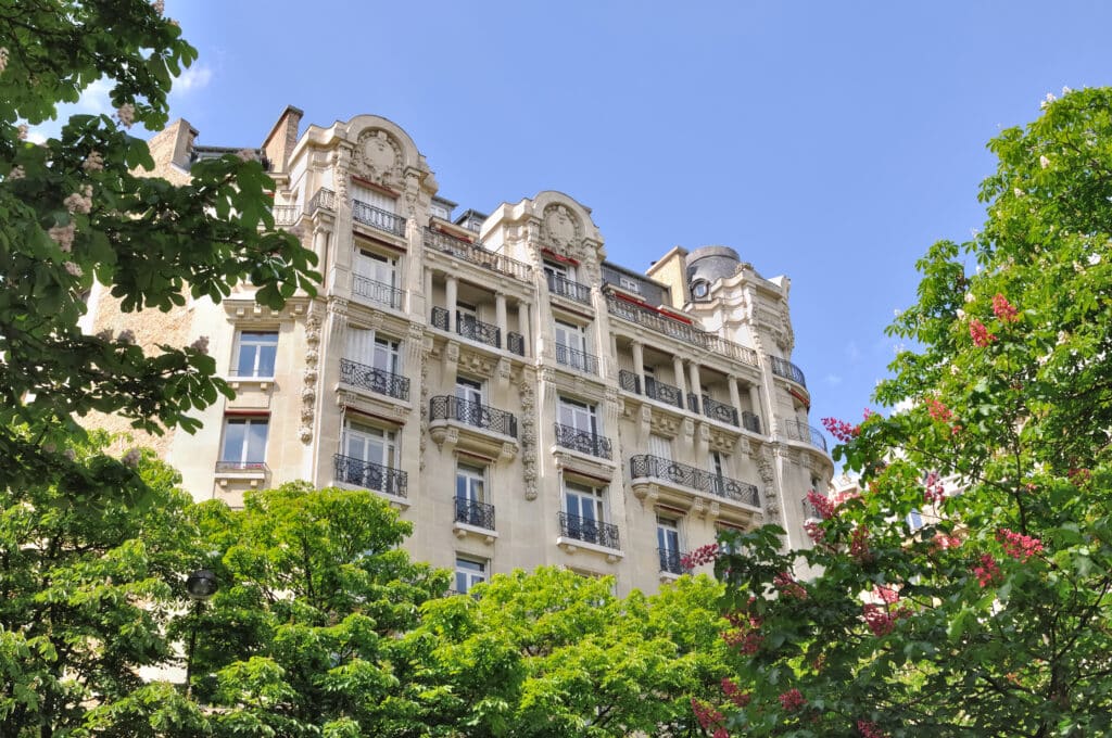 Immeuble parisien orné - Façade typique & arbres fleuris Façade ornée d'un immeuble parisien typique avec balcons en fer forgé, vue à travers des arbres verts fleuris roses et blancs sous un ciel bleu.