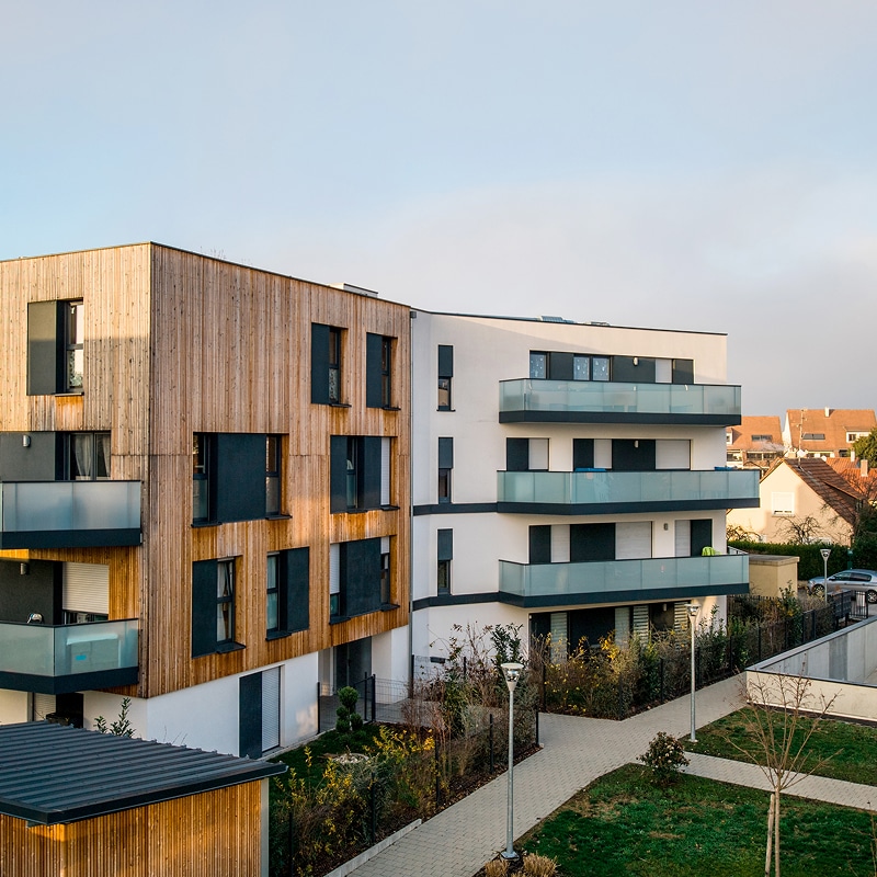 Immeuble résidentiel moderne avec façade en bois et partie blanche aux balcons vitrés. Aménagement paysager devant.