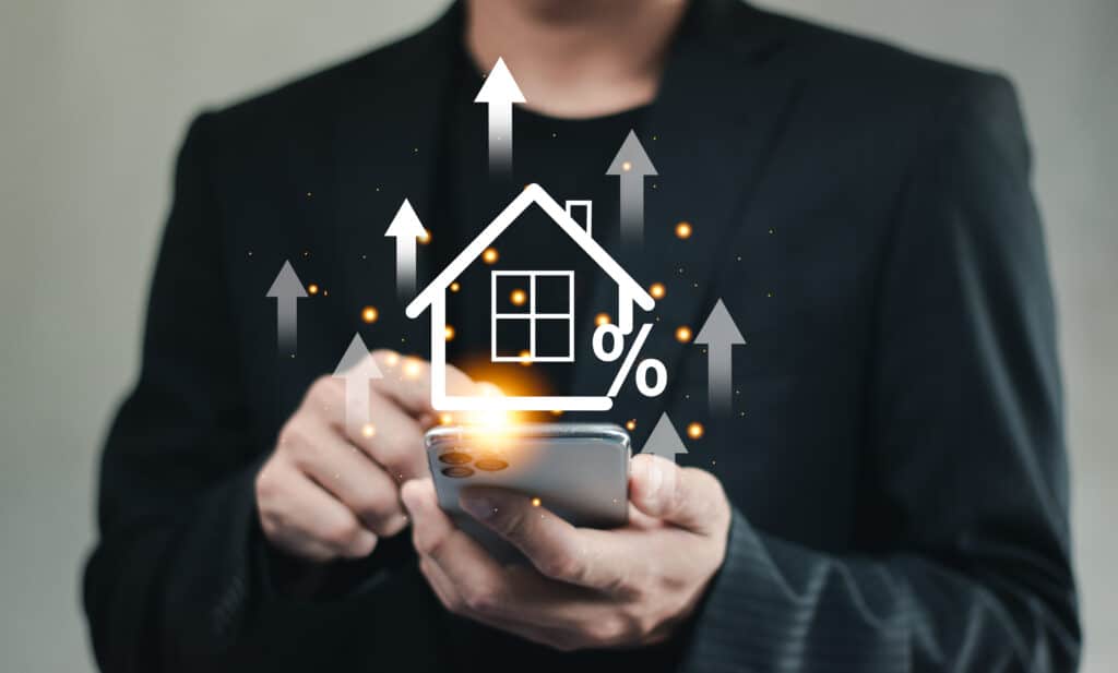 Immobilier Digital : Taux et Valeur en Hausse Homme sur smartphone affiche une icône de maison digitale avec pourcentage et flèches montantes, symbolisant croissance immobilière ou taux.
