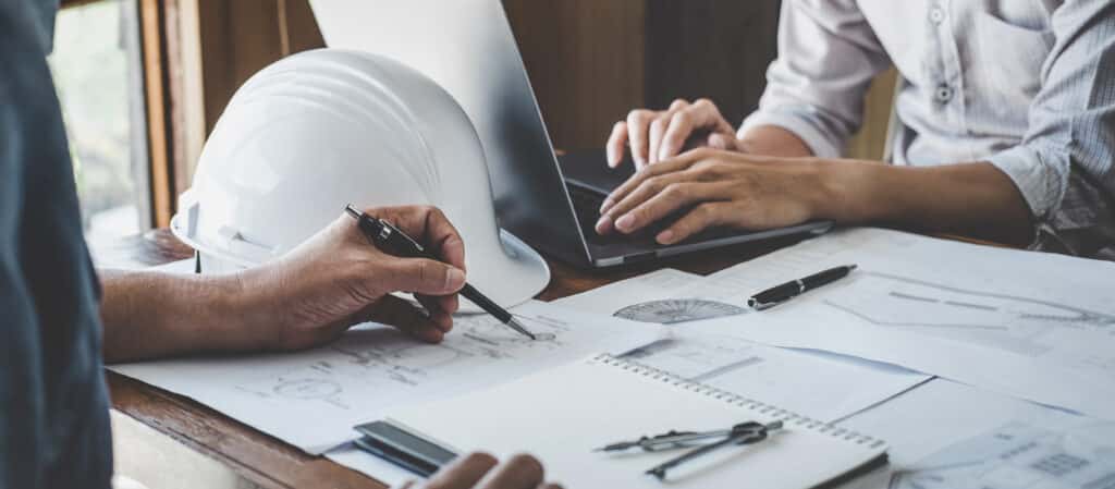 Deux ingénieurs travaillent sur des plans et un ordinateur portable. L'un pointe du doigt, l'autre tape. Casque, plans, outils sur le bureau.