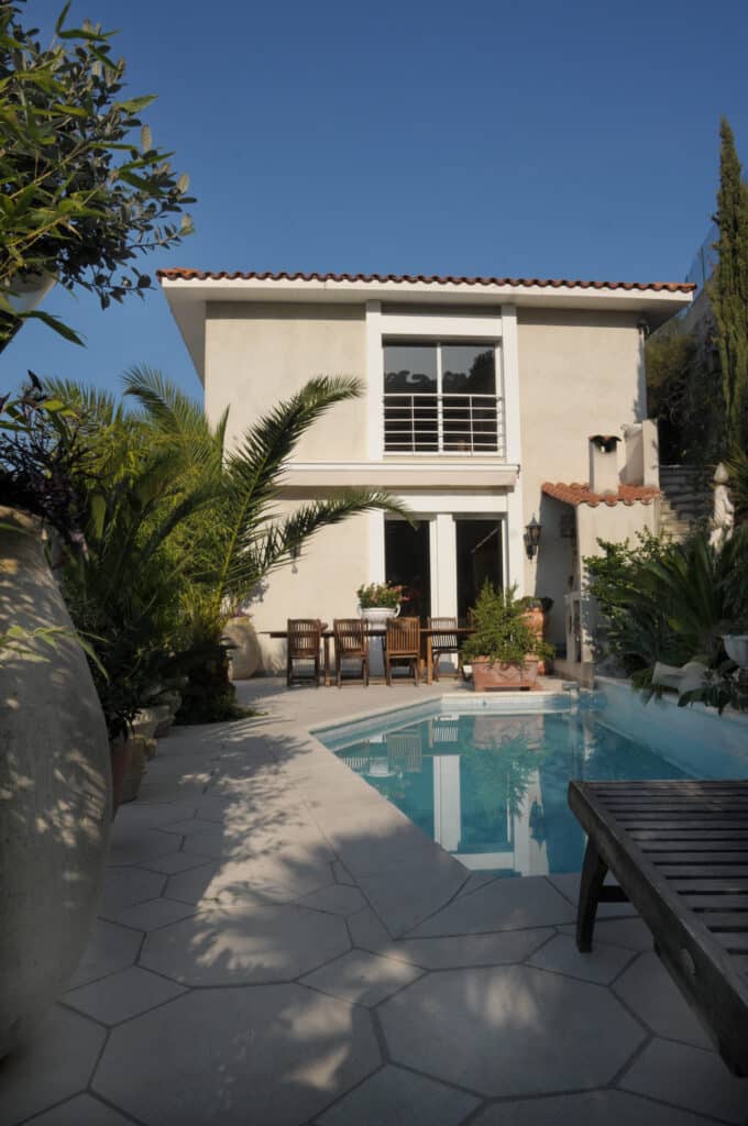 Maison méditerranéenne ensoleillée avec piscine, terrasse, table à manger et végétation luxuriante. Journée claire.