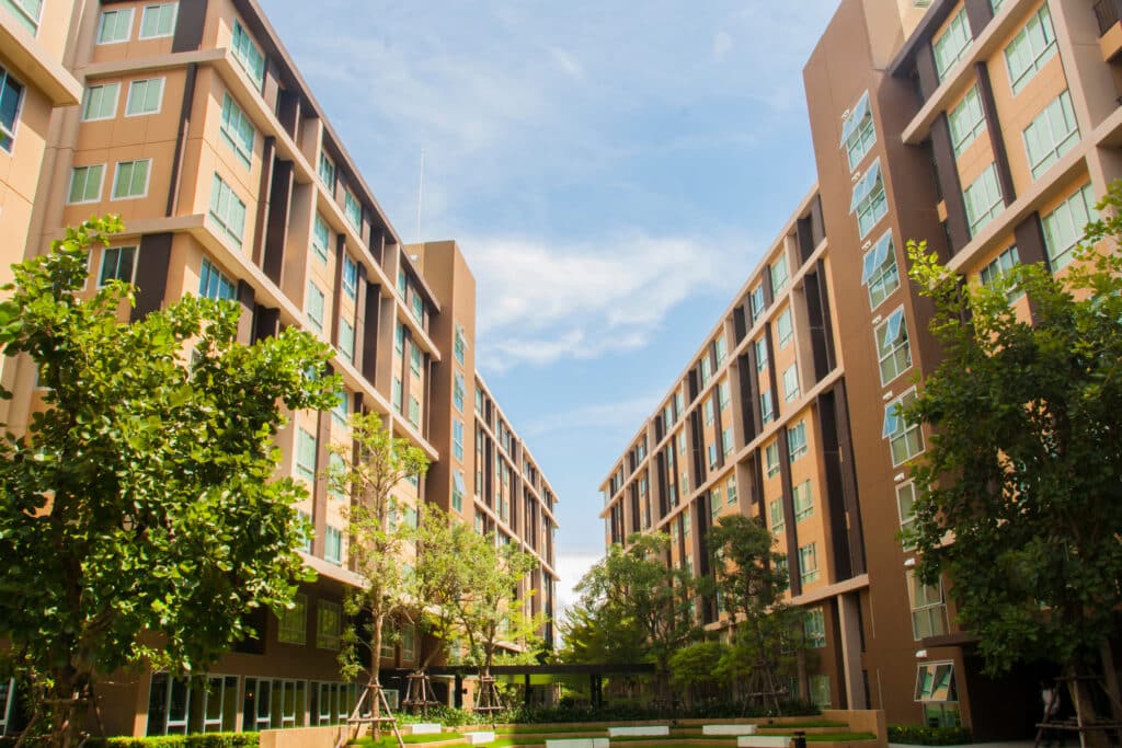 Deux grands immeubles d'appartements bruns font face à un jardin paysagé avec des arbres verts sous un ciel bleu.