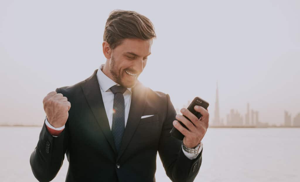 Homme d'affaires en costume sourit, poing levé, regardant son téléphone, exprimant le succès. Skyline de Dubaï.